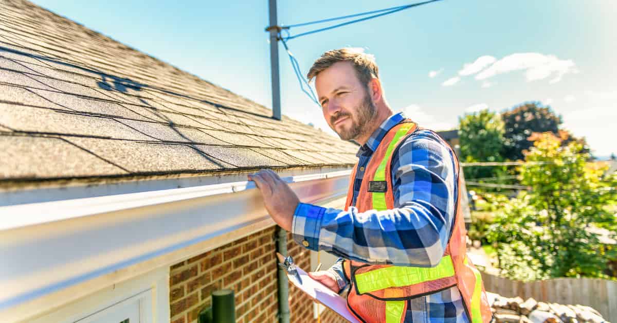 A man in a safety vest examines a house's gutters under a bright sunny sky.